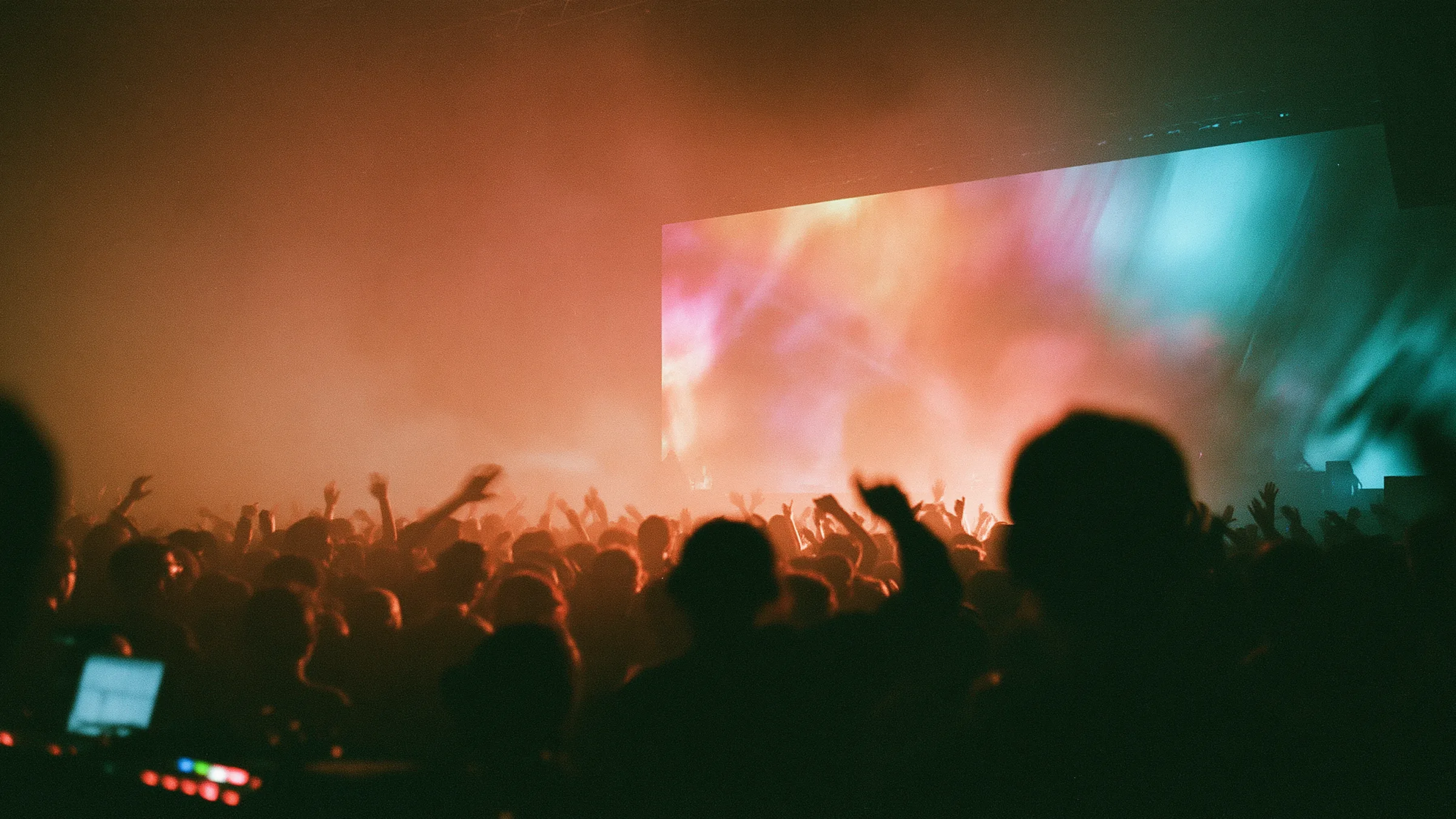 A packed crowd with raised hands facing a glowing video projection screen at a live show